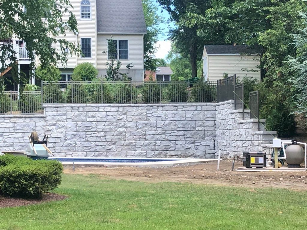 MagnumStone 12 foot retaining wall in a backyard, allowing room for a swimming pool. The wall features an inside corner and top of wall details like caps, fence posts, and step ups.